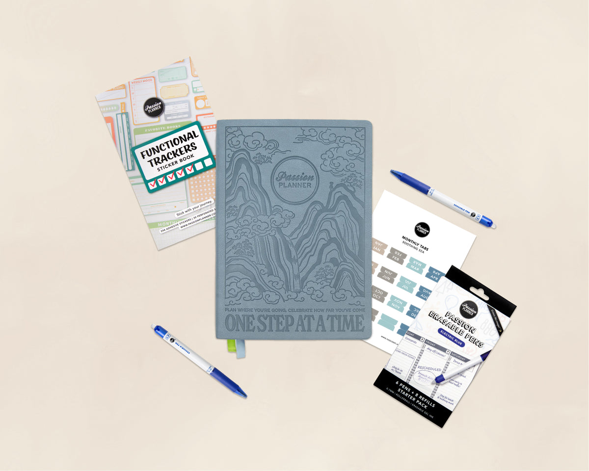A styled flat-lay of a blue-gray embossed planner notebook in the center, surrounded by coordinating planner accessories including functional tracker stickers, blue pens, and monthly tabs on a warm beige background.