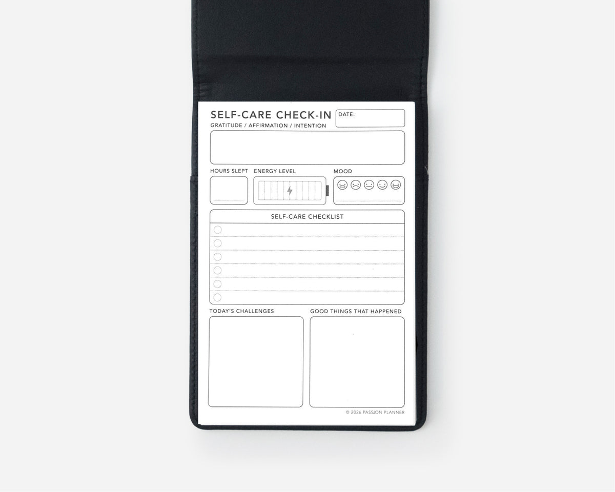 A black self-care checklist notepad shown open against a light gray background, featuring a white insert with fields for date, self-care check-in, sleep, energy level, mood, challenges, and good things that happened.