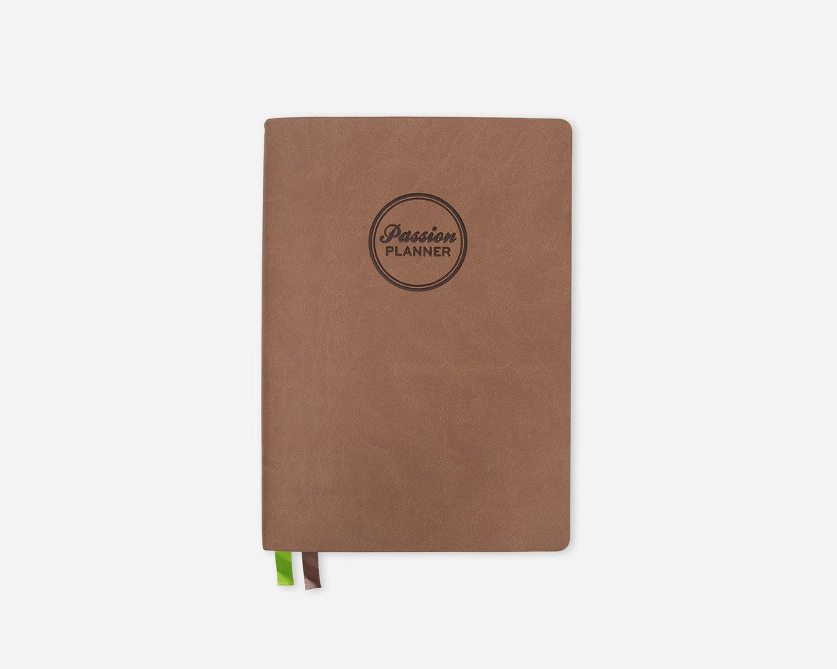 A brown softcover planner with a circular black logo reading 'Passion Planner' on the front, shown on a light gray background with a small green and brown ribbon bookmark peeking out from the bottom.