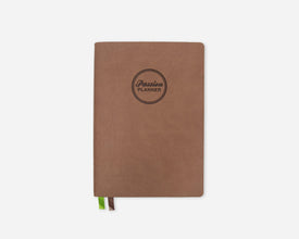 A brown softcover planner with a circular black logo reading 'Passion Planner' on the front, shown on a light gray background with a small green and brown ribbon bookmark peeking out from the bottom.