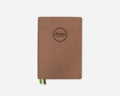 A brown softcover planner with a circular black logo reading 'Passion Planner' on the front, shown on a light gray background with a small green and brown ribbon bookmark peeking out from the bottom.
