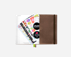 An open brown softcover planner with an elastic closure, shown open with colorful sticker sheets and a lined page fanned out against a light gray background.