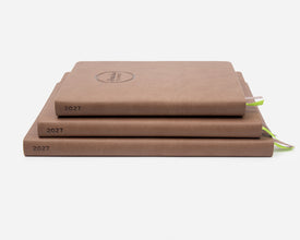 Three stacked hardcover planners in a warm brown, each labeled '2027' on the spine, with small green ribbon bookmarks visible on the right, photographed on a clean white background.