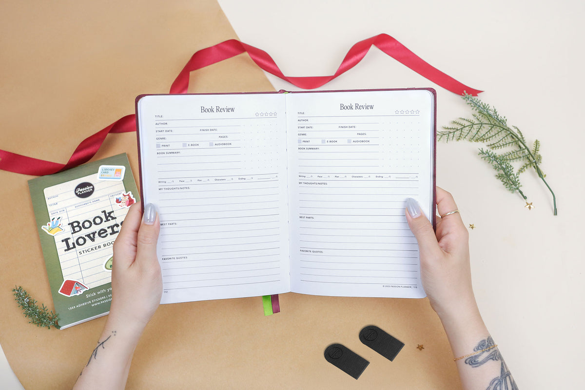 Open reading journal with a book review template on a beige surface with a red ribbon and greenery.