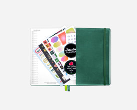 An open green 2027 planner with an elastic closure, shown open with colorful sticker sheets and a lined page fanned out against a light gray background.