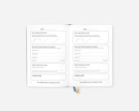 An open Mindful Minute Journal showing two inner pages with lined writing sections and a small blue ribbon bookmark, photographed on a clean light gray background.