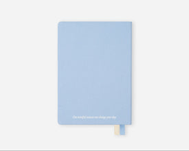 A light blue hardcover journal with a small white quote printed at the bottom and a matching light blue bookmark ribbon, shown on a clean white background.