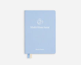 A light blue hardcover journal with a white spiral logo and the text 'Mindful Minute Journal' on the front, shown against a plain white background with a small cream bookmark and blue ribbon peeking out from the bottom.