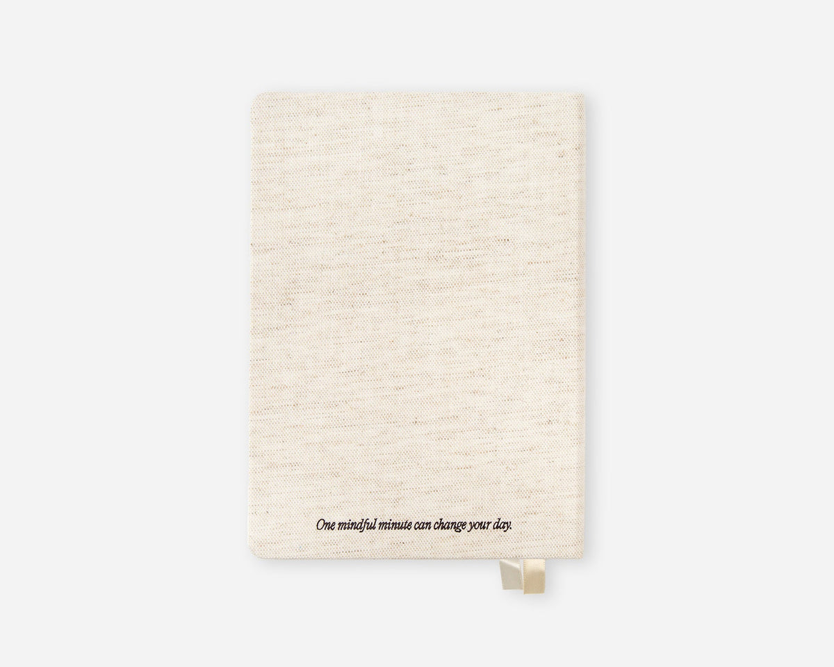 A cream-colored linen-textured notebook with a small gold bookmark and the printed quote 'One mindful minute can change your day.' at the bottom, shown on a light gray background.