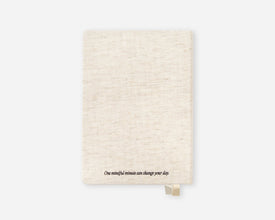 A cream-colored linen-textured notebook with a small gold bookmark and the printed quote 'One mindful minute can change your day.' at the bottom, shown on a light gray background.