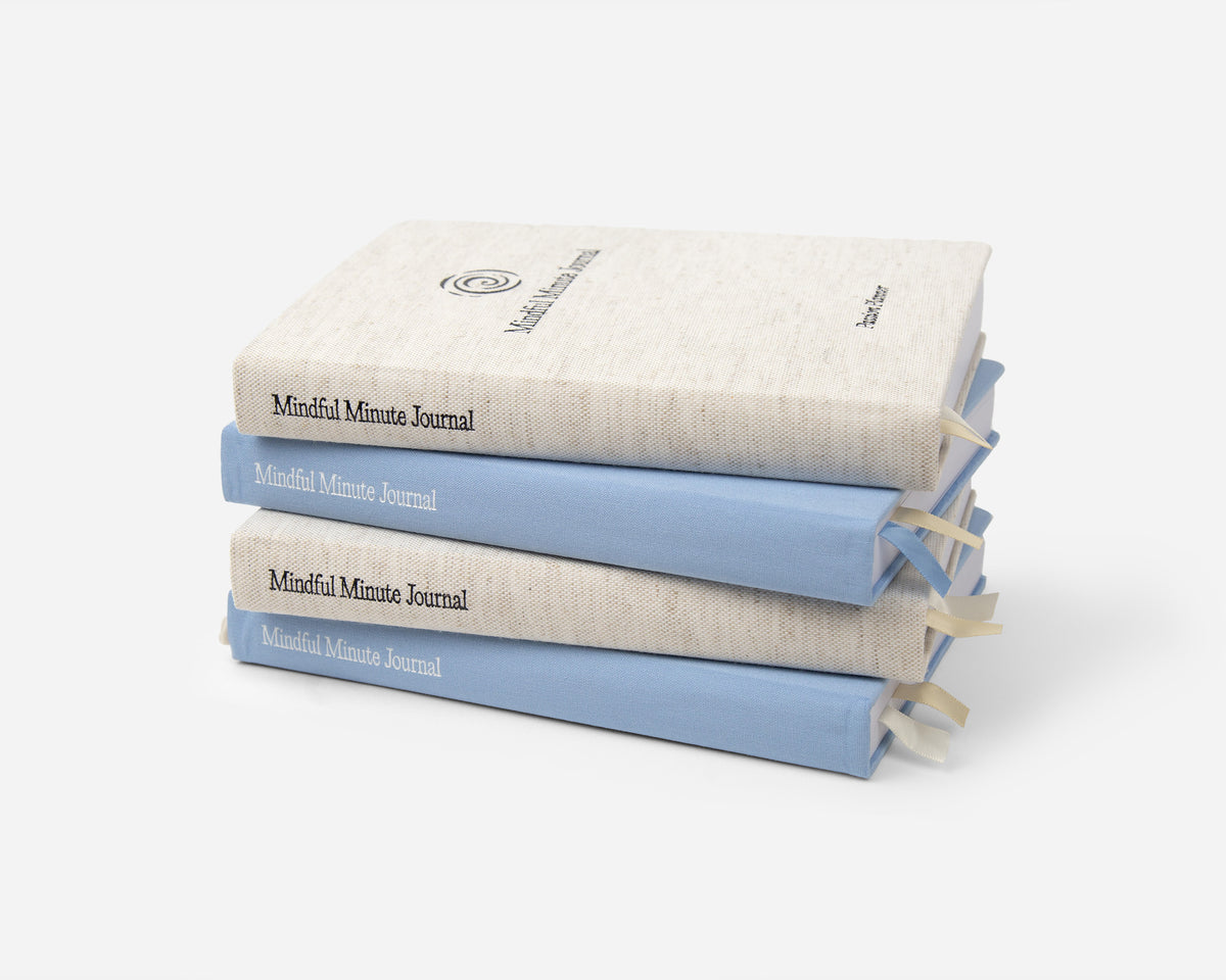 A stack of 4 Mindful Minute Journals in linen and blue on a gray background.