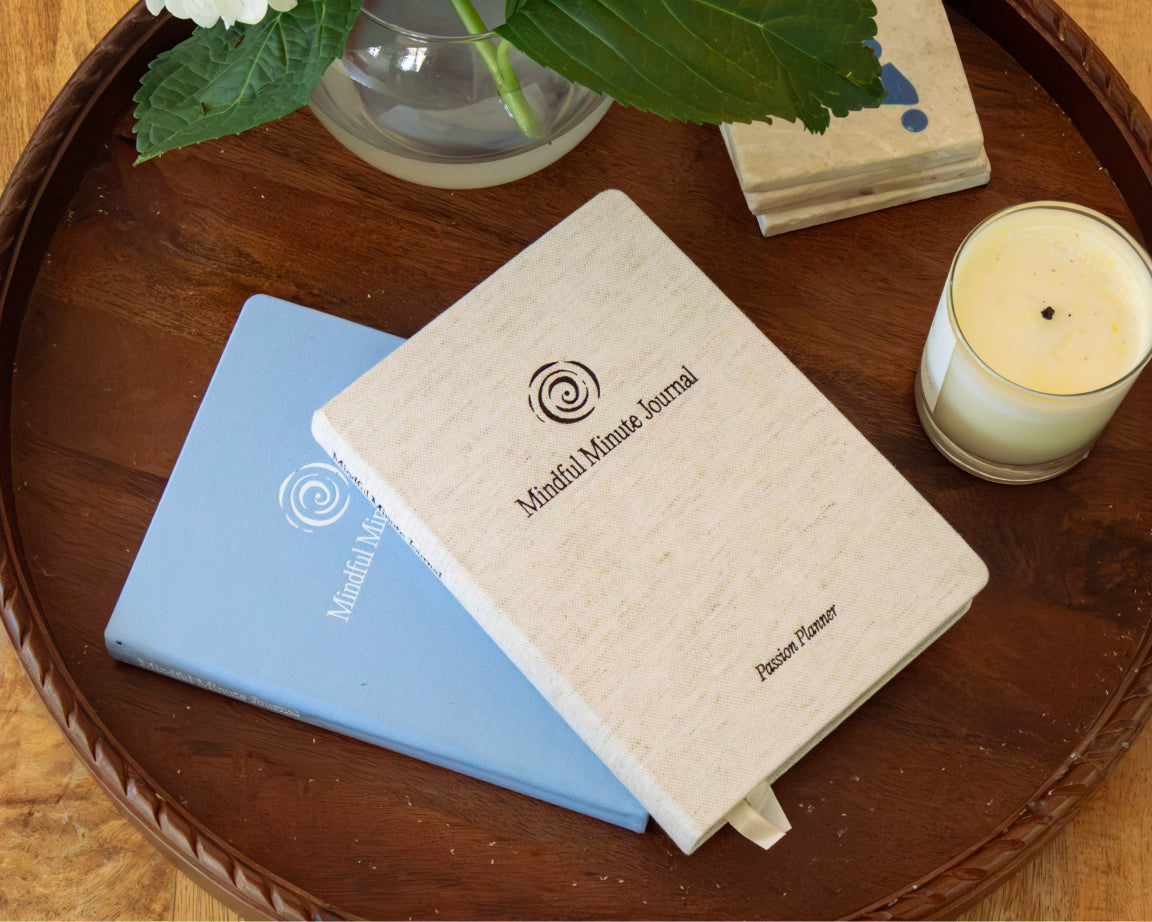Two 'Mindful Minute Journal' notebooks, one in natural beige and one in light blue, arranged on a round wooden tray alongside a candle, flowers, and coasters.