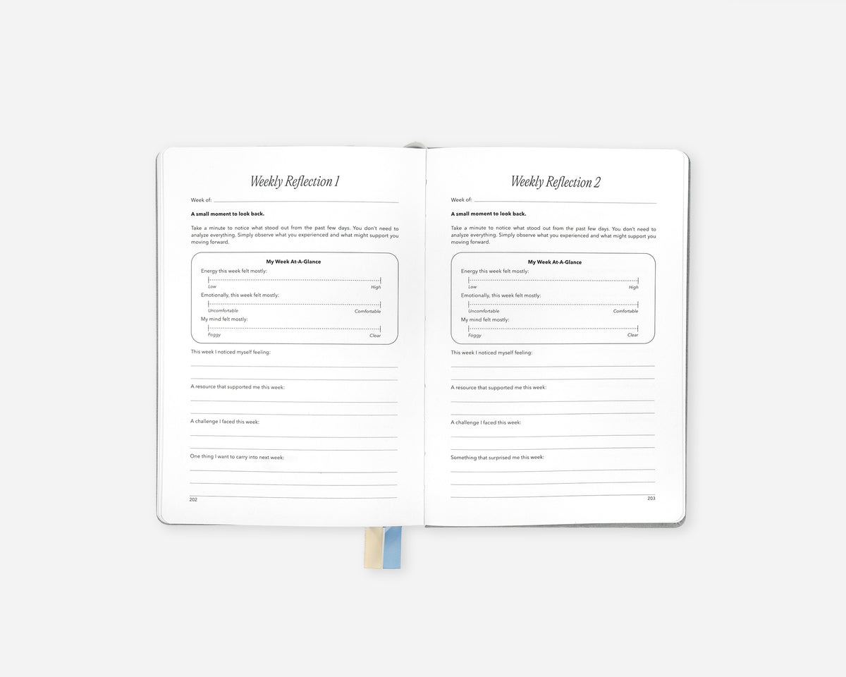 An open Mindful Minute Journal showing two reflection pages with lined writing areas and prompts titled 'Weekly Reflection 1' and 'Weekly Reflection 2'. A small blue ribbon bookmark is visible at the bottom center against a light gray background.
