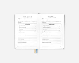 An open Mindful Minute Journal showing two reflection pages with lined writing areas and prompts titled 'Weekly Reflection 1' and 'Weekly Reflection 2'. A small blue ribbon bookmark is visible at the bottom center against a light gray background.