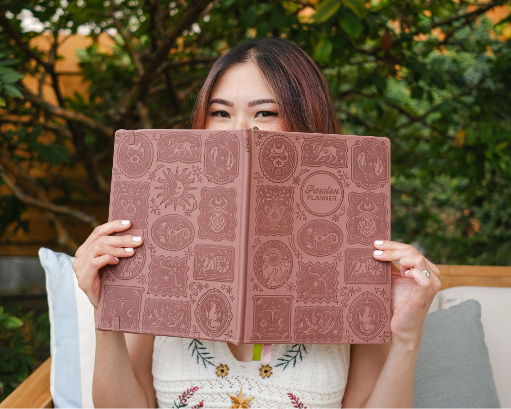 A woman holding a large embossed planner in front of her face, with a visible &