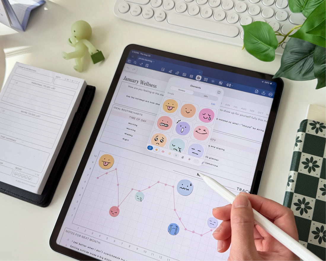 A hand holding a white stylus points at a tablet screen showing a wellness tracking digital planner dashboard. The tablet sits on a white desk surrounded by a notebook, a small green figurine, a keyboard, and a plant.