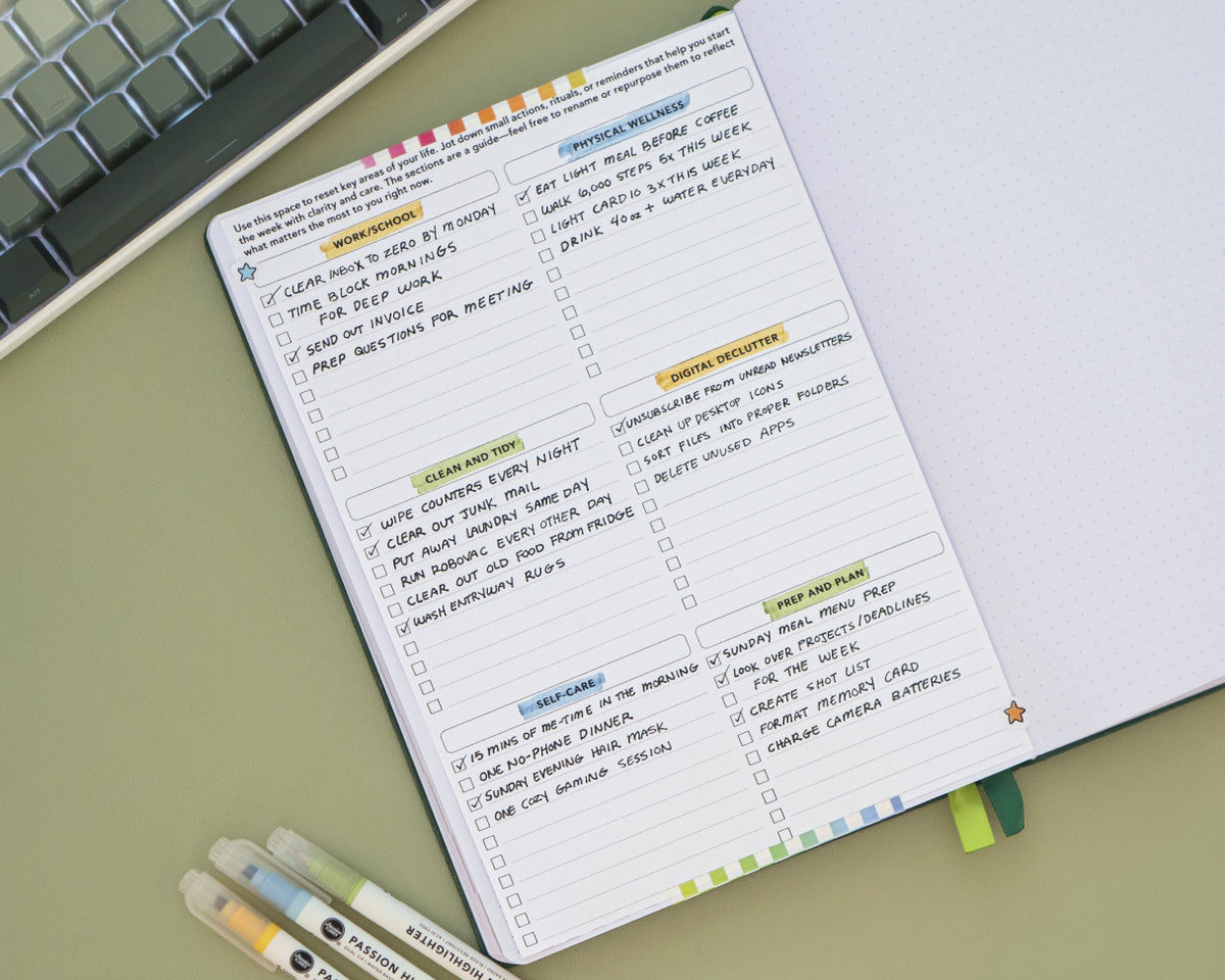 An open planner or notebook showing a weekly reset planner page with colorful section headers and checklist boxes, photographed on a light green surface with part of a keyboard and pens visible.